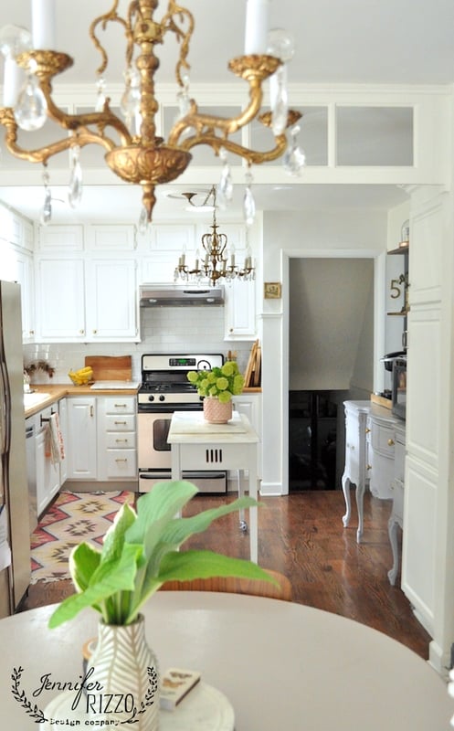 Bright kitchen with white cabinets, stainless steel appliances, wooden floors, a central island with green flowers, and gold chandeliers. Kitchen subway tile adds a timeless touch behind the counters, while a potted plant sits in the foreground.