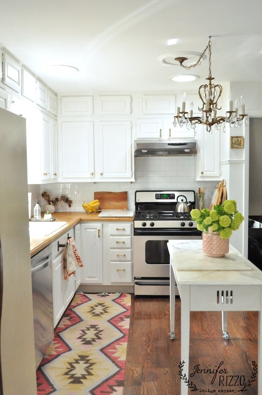 A bright kitchen with white cabinets, stainless steel appliances, classic kitchen subway tile, a small island topped with a pink vase of green flowers, a patterned rug, and a chandelier.