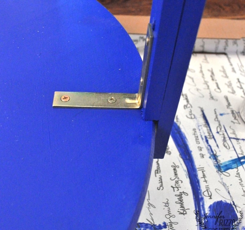 A close-up of a brass L-bracket attaching a blue wooden shelf to a blue vertical support, with handwritten text visible underneath—perfect for your next DIY Penny tile table top project.