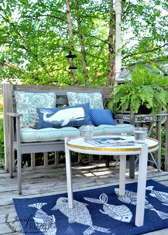 Wooden outdoor bench with patterned cushions and pillows, next to a fern plant. A round white table with a DIY penny tile table top, set with glasses and books, sits on a blue rug featuring whale illustrations on the wooden deck.