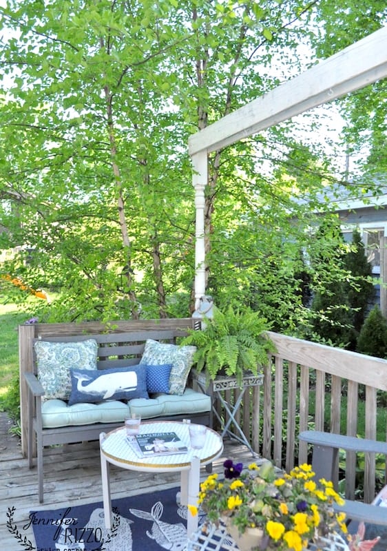 A wooden patio with a cushioned bench, potted flowers, lush green trees in the background, and a small table featuring a stylish DIY penny tile table top adorned with books.