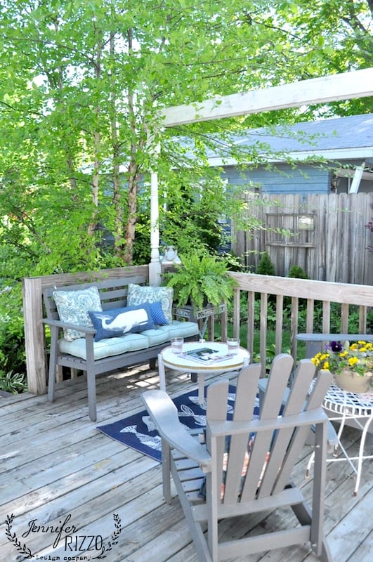 Wooden deck patio with cushioned bench, two chairs, small tables featuring a DIY penny tile table top, potted plants, and a blue rug—surrounded by greenery and a wooden fence.