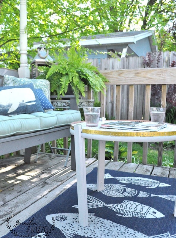Outdoor patio with a wooden bench, two glasses of water on a DIY penny tile table top, and a fish-patterned rug on a weathered deck. Lush green plants and a wooden fence create a serene backdrop.