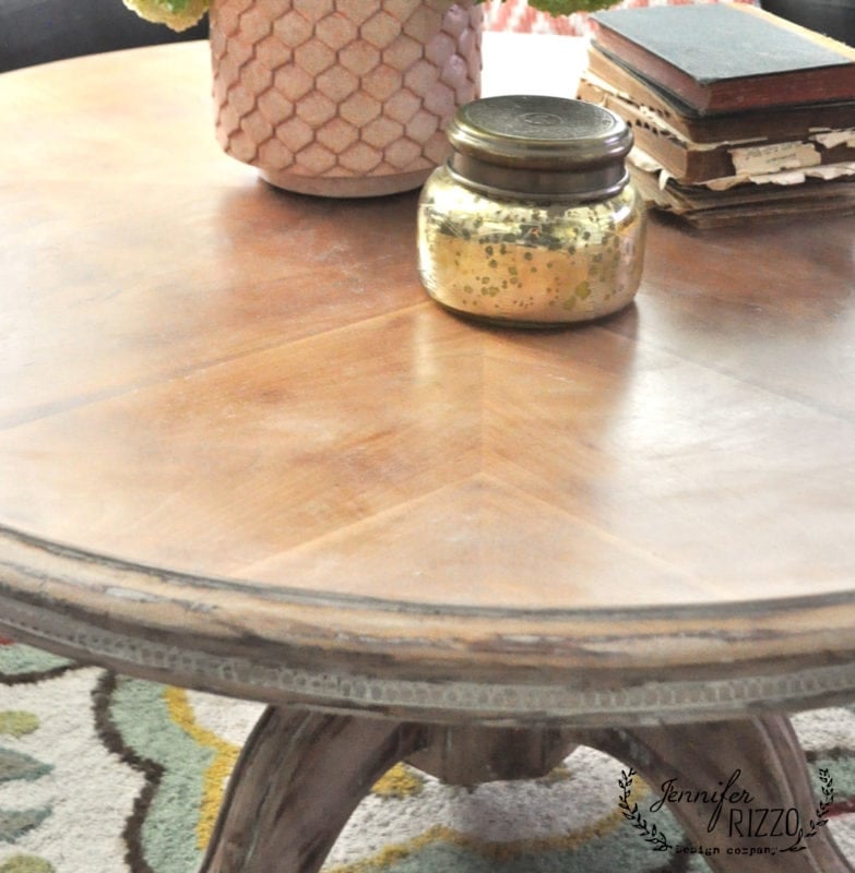 A wood coffee table with white wax finish features a pink textured vase, a closed mason jar, and stacked books on top, all set on a patterned rug.