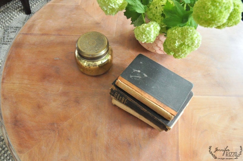 A stack of vintage books, a gold jar, and a vase with green hydrangeas are arranged on a wood coffee table with white wax.