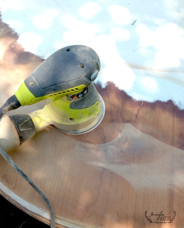 A handheld electric sander is being used to sand the surface of a wood coffee table with white wax, removing the finish and exposing the bare wood.