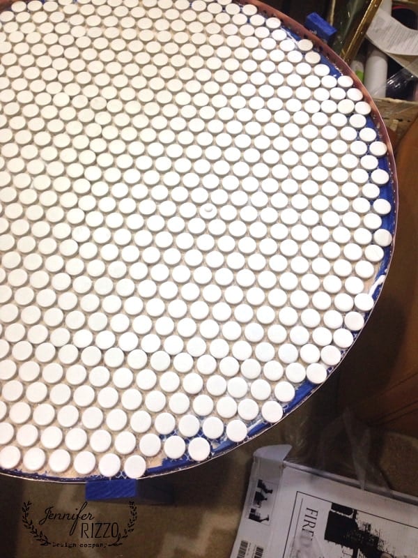 A round DIY penny tile table top covered with a grid of small, white circular tiles; crafting materials and papers are visible nearby.