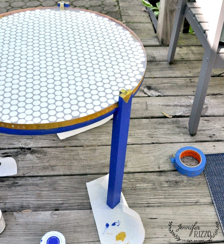 A round table with a blue frame and a white hexagonal-tiled top—perfect for a DIY penny tile table top project—rests on a wooden deck, surrounded by painter's tape and masking paper from the painting process.