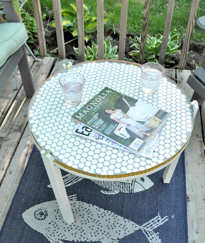 A round outdoor table with a white DIY penny tile table top and hexagon pattern holds two drinking glasses and a Magnolia magazine, set on a fish-patterned rug on a wooden deck.