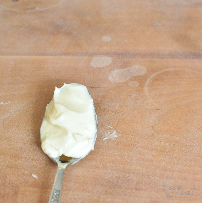 A spoon with a dollop of white cream or mayonnaise rests on a wooden surface, which shows faint rings from furniture.