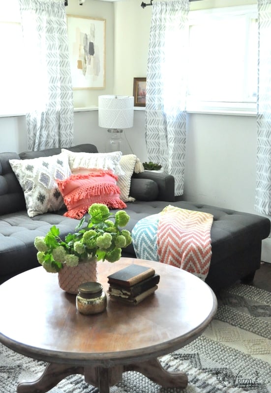 A modern gray and coral family room featuring a dark gray sectional sofa, patterned pillows, a throw blanket, a round wooden coffee table, green plants, and books. Light curtains softly cover the windows.