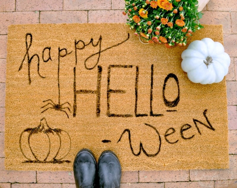 A doormat with the text "Happy HELLO-ween" written in playful script is adorned with drawings of a spider and pumpkins. Nearby, a white pumpkin, a pair of black boots, and a pot of orange flowers hint at the festive October vibes and seasonal decor!