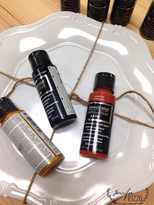 Three bottles of Americana multi-surface satin paint are tied with twine on a white plate with a wooden background, perfect for creating hand lettered Halloween plates.