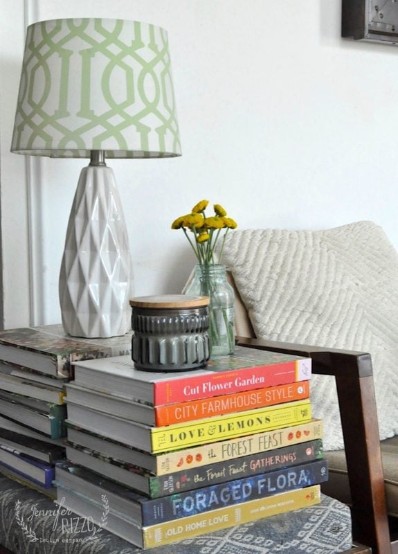 A white geometric table lamp, a small vase with yellow flowers, and a grey candle sit on a stack of books next to a textured pillow on a wooden chair.