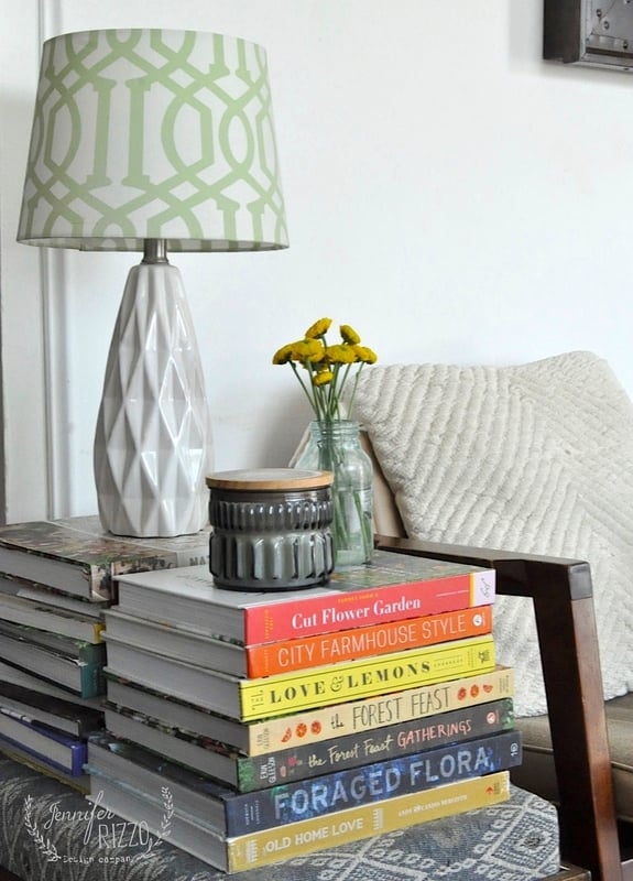A white geometric table lamp, a small vase with yellow flowers, and a grey candle sit on a stack of books next to a textured pillow on a wooden chair.