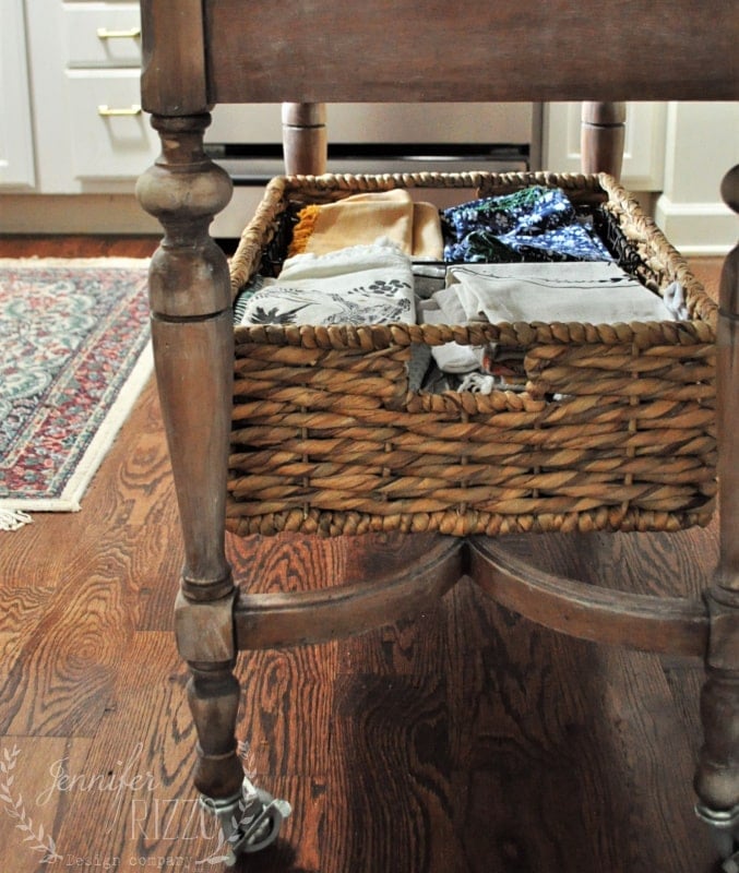 A woven basket filled with folded linens sits on a wooden shelf beneath a table in a kitchen with hardwood floors, adding to the inviting decor with plants arranged nearby.