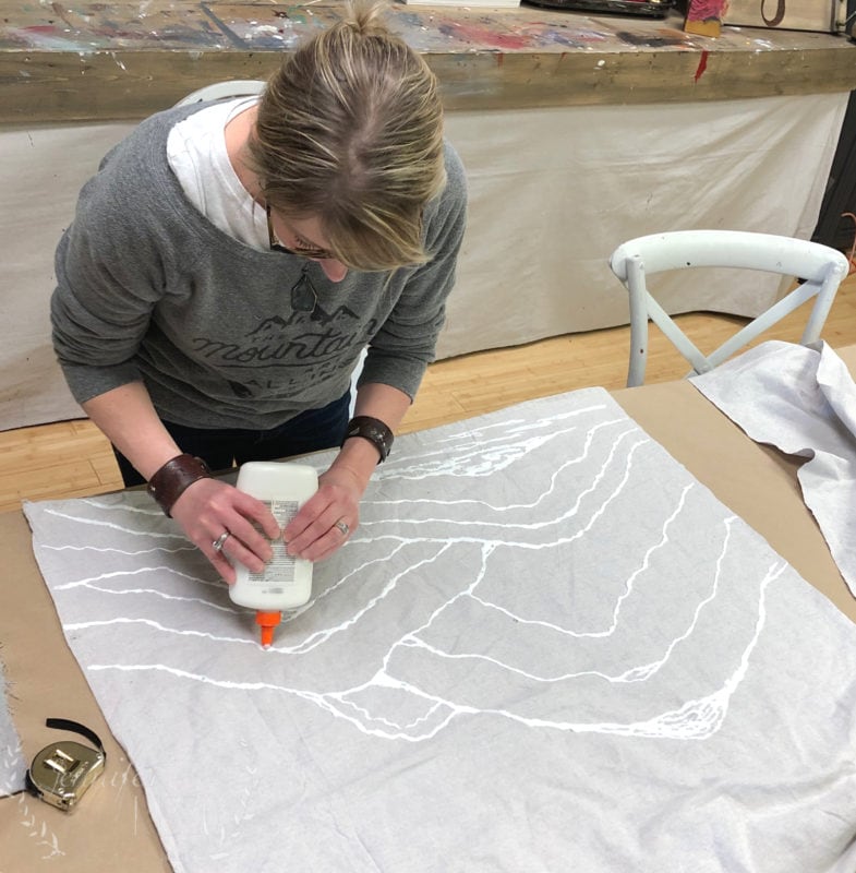 A person is creating glue resist art by applying white glue in patterns on a large piece of fabric laid out on a table in an art studio.