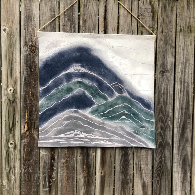 A square textile wall art piece with layered, wavy patterns in shades of blue, gray, and white—resembling mountains—showcases glue resist art as it hangs on a wooden fence by a string.