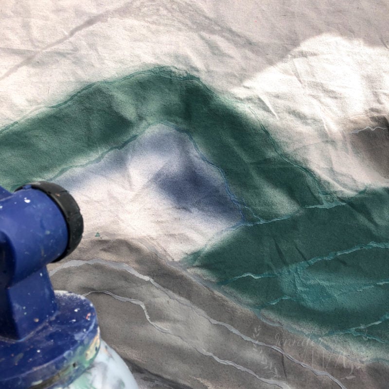 A close-up of fabric being spray painted with green and blue colors using glue resist art, with part of a blue spray paint can visible in the corner.