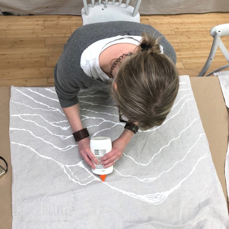 A person applies glue in wavy lines onto a piece of fabric laid out on a table, using a white plastic bottle, creating patterns for glue resist art.