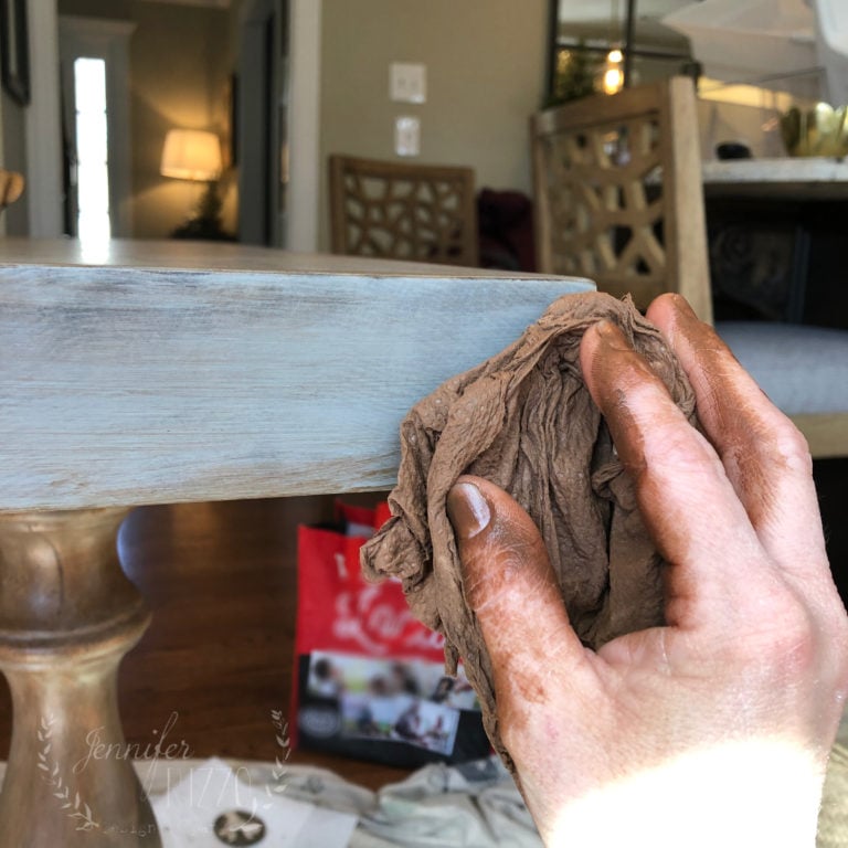A person with stained hands uses a cloth to wipe the edge of a wooden table in a well-lit room after a Painted Kitchen Table Makeover.