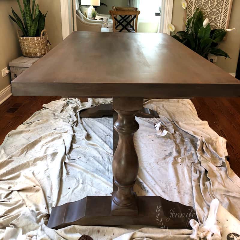 A wooden dining table with a pedestal base, likely undergoing a Painted Kitchen Table Makeover, is placed on a drop cloth in a room with hardwood floors and neutral decor.