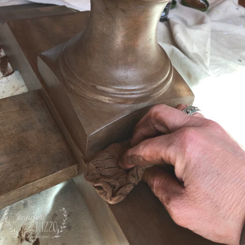 A hand uses a cloth to wipe or stain a wooden pedestal with a brown finish, perfect for your next Painted Kitchen Table Makeover.