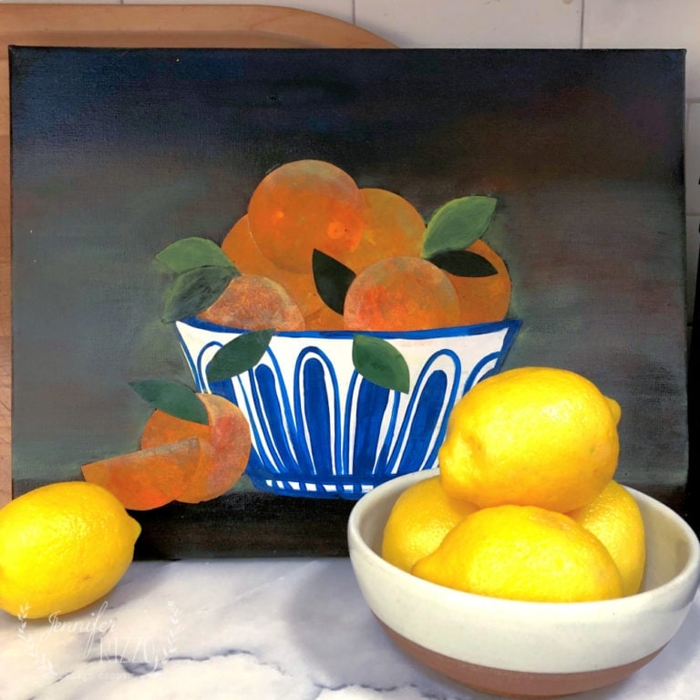 A still life painting of oranges in a blue and white bowl sits behind a real bowl filled with three yellow lemons, with two additional lemons placed beside it.