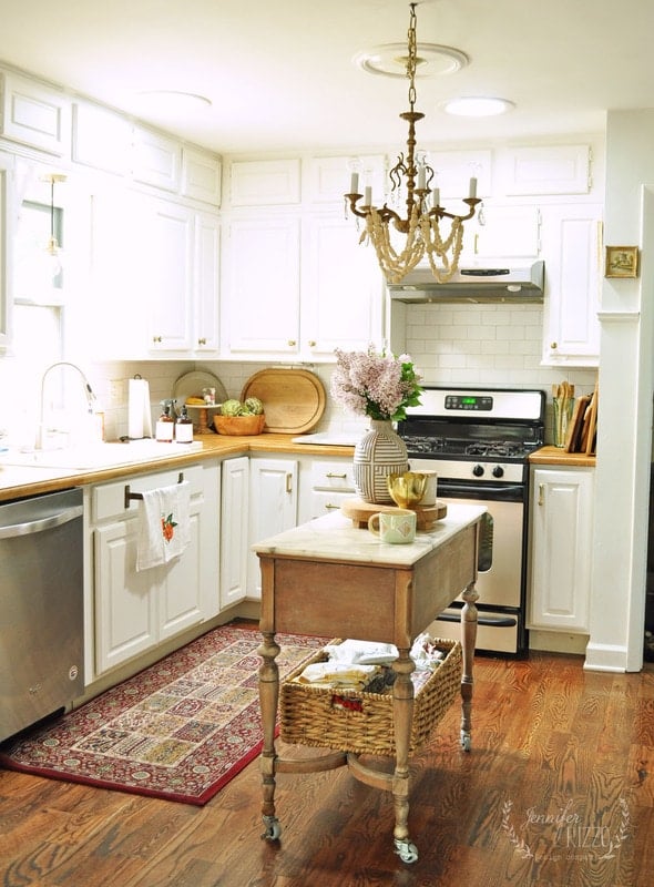 Bright kitchen with white cabinets, a small wooden island with a basket shelf, stainless steel appliances, a decorative rug, and a chandelier hanging from the ceiling—plus a charming Create A Cut Paper Decoupage Orange Still Life Canvas on display.