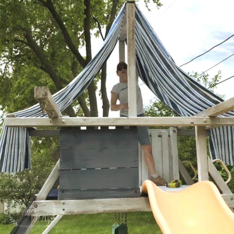 Person standing on a wooden playset structure with a new fabric canopy draped as a striped cover and a yellow slide in the backyard.