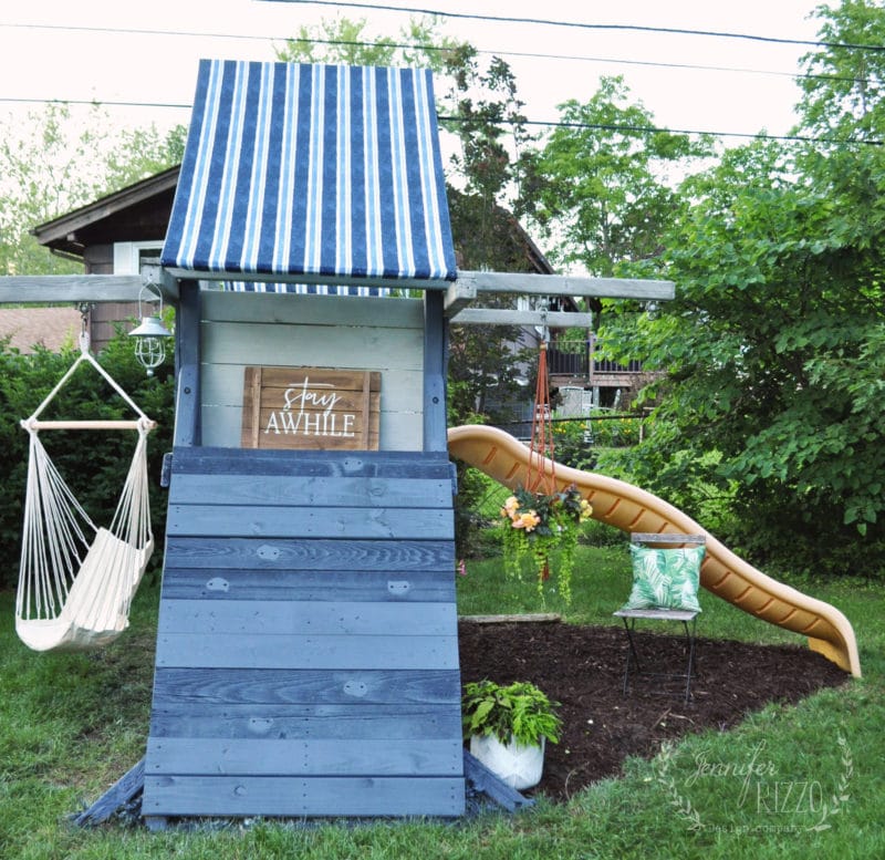 A wooden play structure with a new fabric canopy for a playset, blue striped, a climbing wall, beige slide, hanging chair, and green metal chair in a grassy yard.