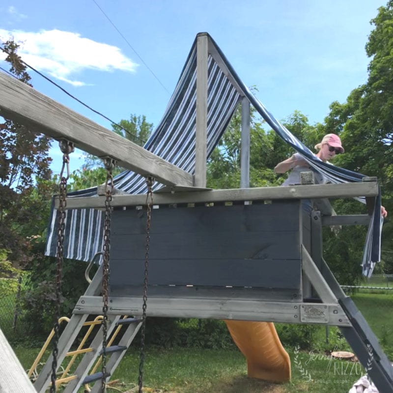 Person installing a new fabric canopy for a playset, featuring striped material on a wooden structure in a backyard surrounded by trees and a blue sky.