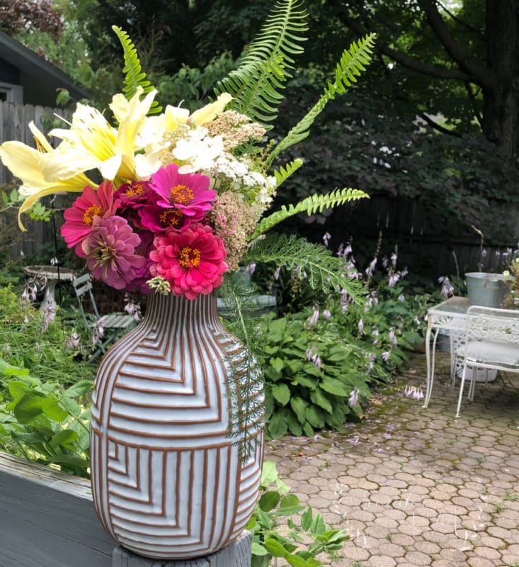 A geometric-patterned vase holds a bouquet of assorted flowers and ferns, placed on a wooden surface in a garden with greenery—perfect inspiration to make backyard bouquets—with patio furniture in the background.