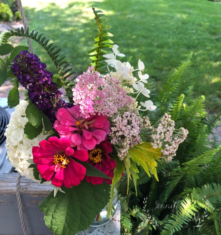 A mason jar filled with assorted cut flowers, including pink and white blooms, purple flowers, and green foliage, showcases how you can make backyard bouquets, displayed outdoors on a wooden surface with grass in the background.