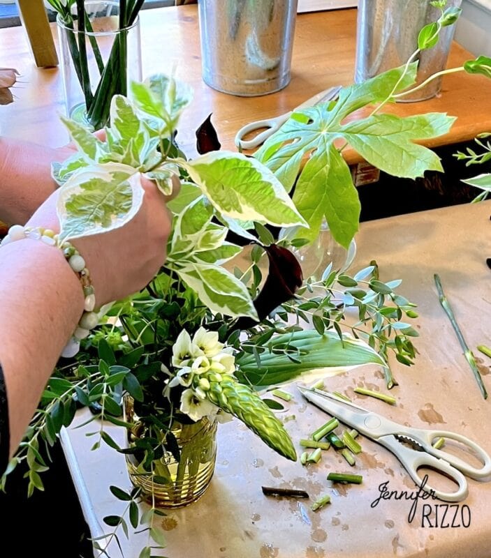 A person creates backyard bouquets, arranging green and white variegated leaves and flowers in a gold vase on a table with scissors and plant stems.