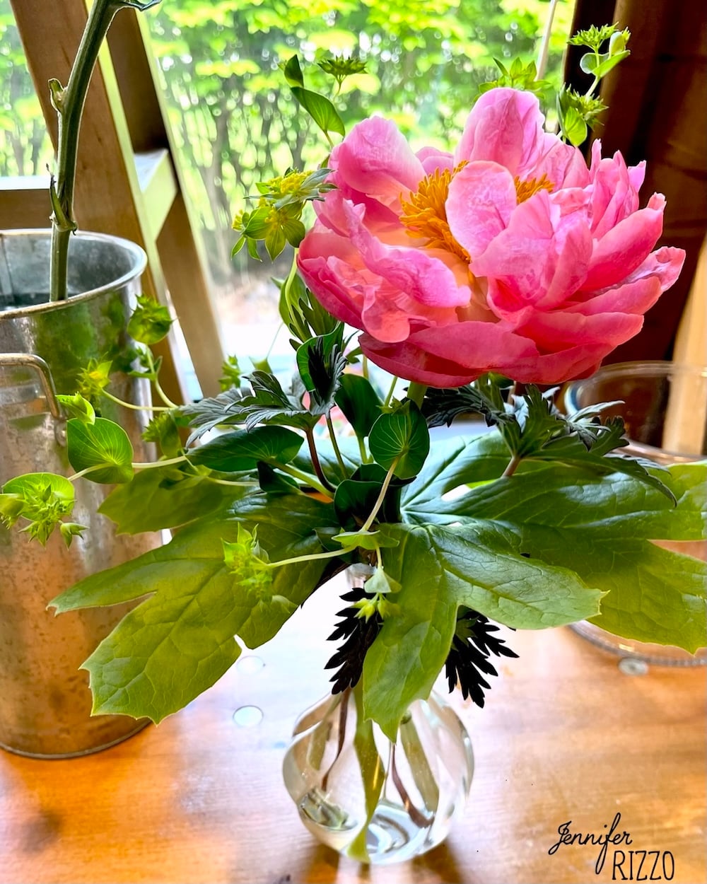A pink peony with green leaves in a small glass vase sits on a wooden surface near a window, inspiring you when creating flower bouquets for gifting to make backyard bouquets, with a metal container and greenery in the background.