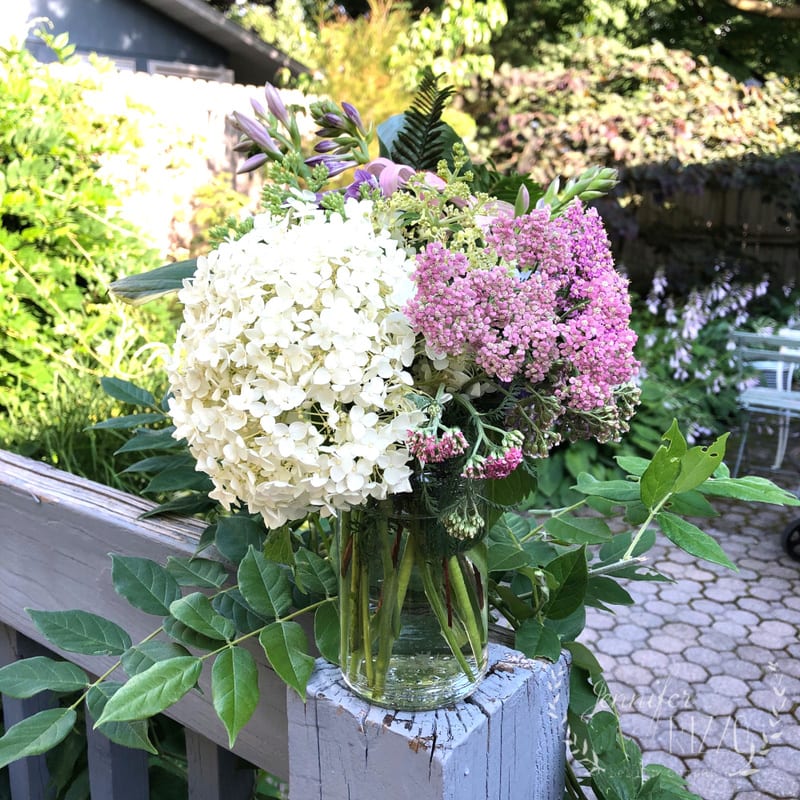A glass jar filled with white and pink flowers sits on a wooden railing in a sunlit garden, inspiring you to make backyard bouquets amid lush greenery.