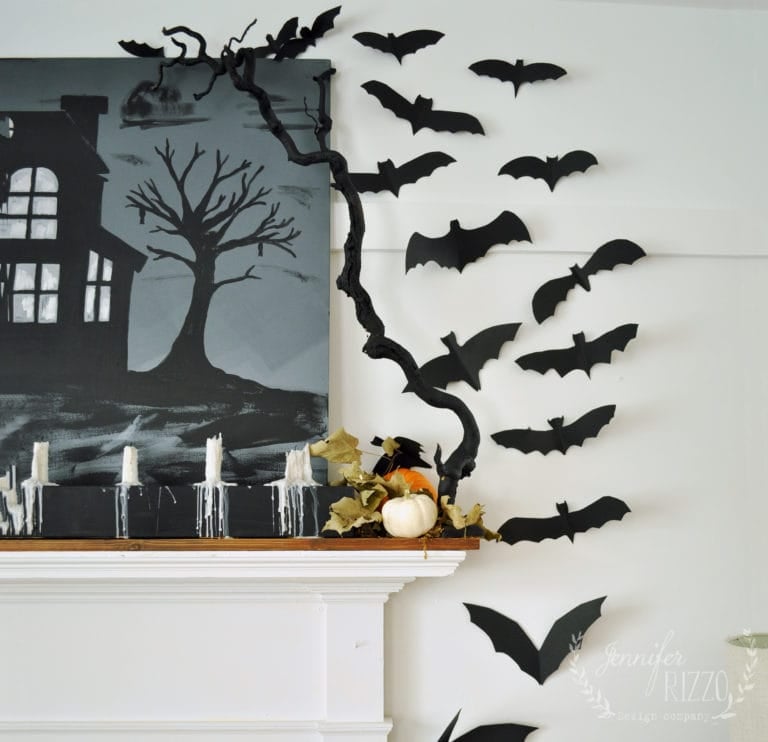 Black paper bats and a branch are arranged above a mantel with a DIY drippy mantel candelabra, candles, a pumpkin, and a painting of a haunted house, creating an eerie Halloween decoration.