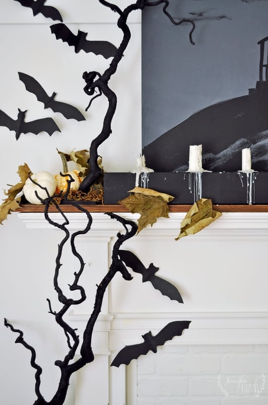 A white mantel decorated with black paper bats, dried leaves, white pumpkins, and a DIY drippy mantel candelabra alongside white candles, creating a Halloween-themed display.