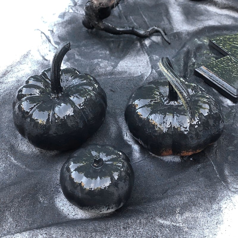 Three small pumpkins coated in shiny black paint sit beneath a spray painted spooky tree, all resting on a surface dusted with black and silver spray paint for striking Halloween decor.
