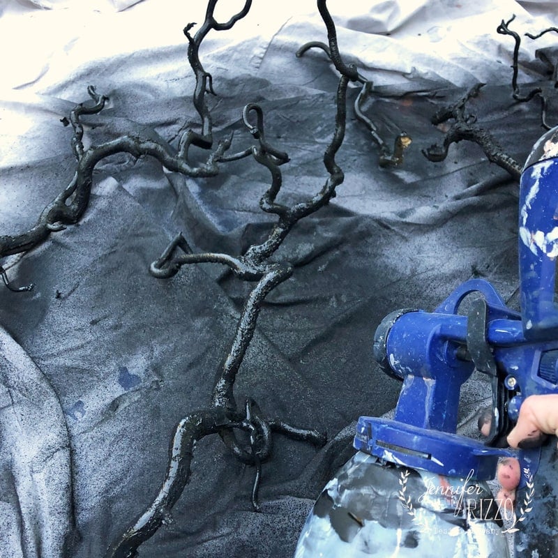 A person uses a blue paint sprayer to coat twisted branches black for spray painted spooky tree Halloween decor, with a tarp underneath to catch overspray.