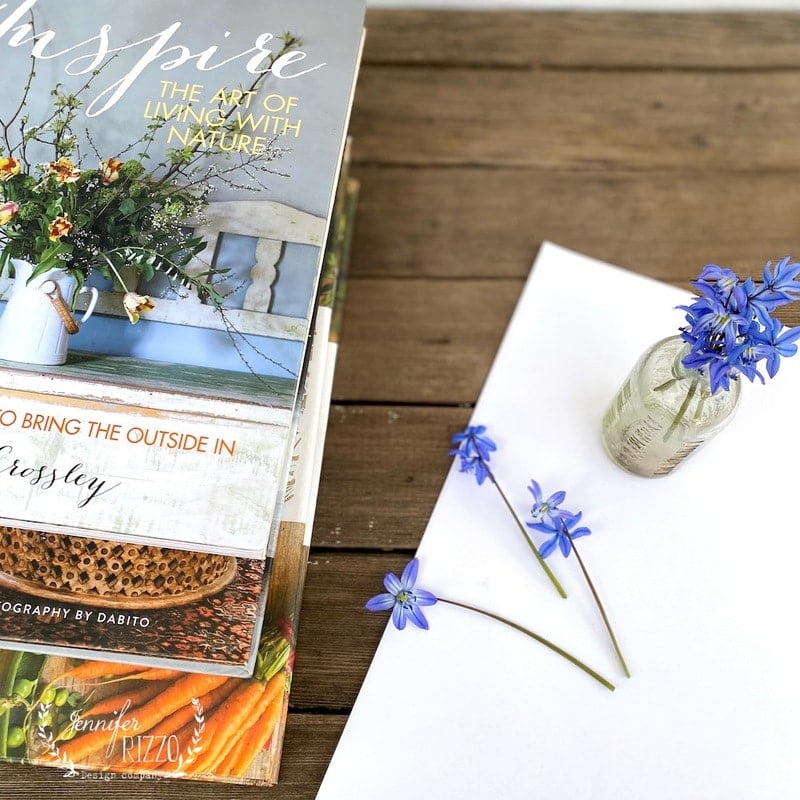 A book titled "The Art of Living with Nature," perfect for learning how to press flowers with books, sits on a table beside a blank sheet of paper and a small glass jar filled with blue flowers.