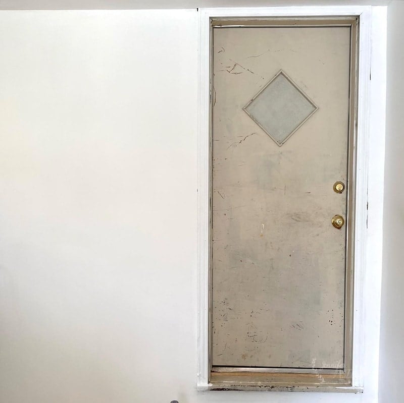 A beige door with a diamond-shaped window is set into a white wall, where mudding and taping the drywall has left a smooth finish, slightly elevated above the floor with no visible steps or landing.