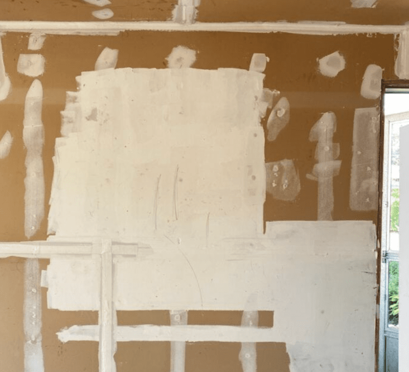 Room interior with unfinished drywall, visible joint compound patches from mudding and taping the drywall, and an open door leading outside. Walls show areas prepared for painting or further construction work.