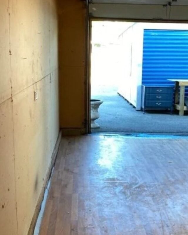 An empty garage with wooden flooring, an open garage door, and outdoor storage units visible in the background—perfect for mudding and taping the drywall to finish your project.