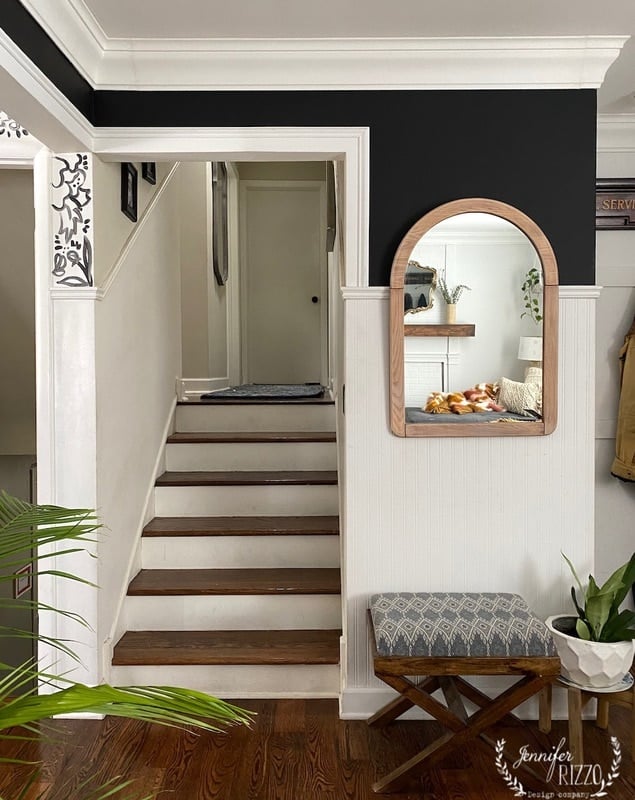 A staircase leads up to a closed white door; a wall mirror reflects a living room with a couch. A bench with a cushion and plant are in the foreground.