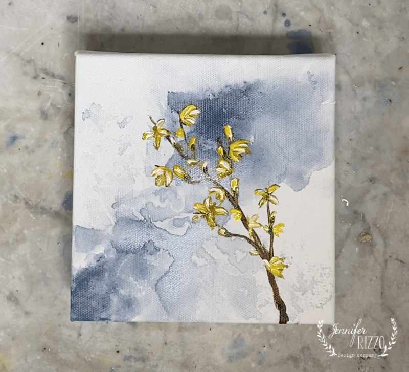 A small canvas painting features a brown branch with yellow flowers against a blue and white watercolor background, offering inspiration for those learning how to paint a forsythia branch.