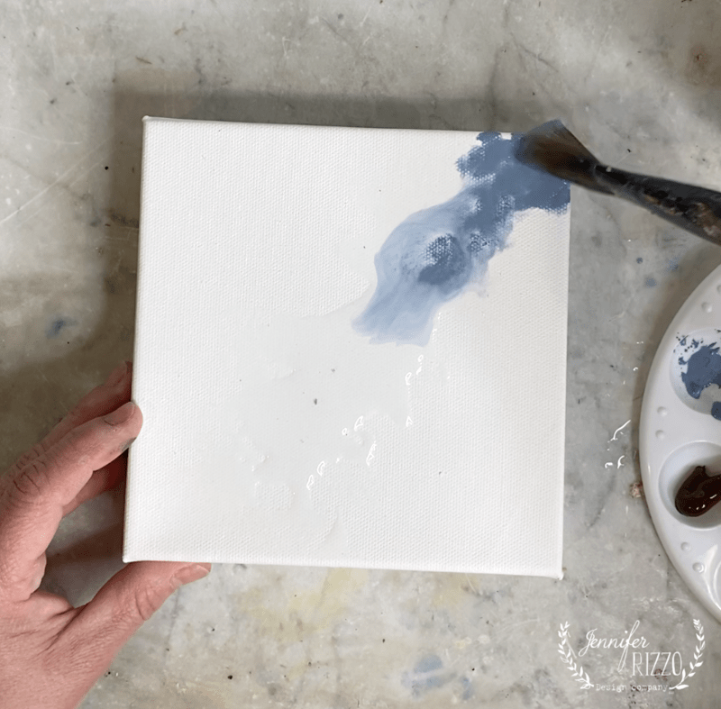 A hand holds a blank white canvas while a paintbrush applies blue paint to the upper right corner; nearby, a white palette with paint is partially visible—perfect for learning how to paint a forsythia branch.