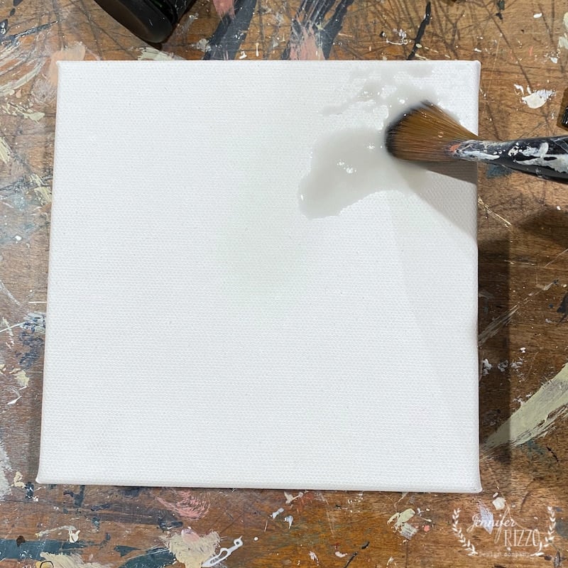 A paintbrush applies a clear liquid to a blank white canvas resting on a paint-splattered work surface, preparing the base for learning how to paint a forsythia branch.