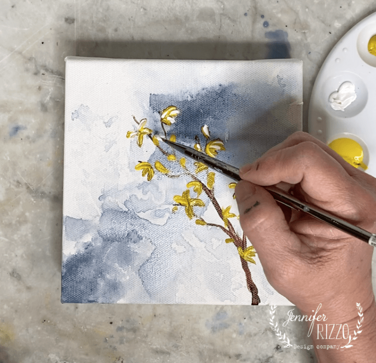 A hand is painting yellow flowers, demonstrating how to paint a forsythia branch on a small canvas with a brush. A palette with yellow and white paint sits nearby, and the canvas features a blue and white background.